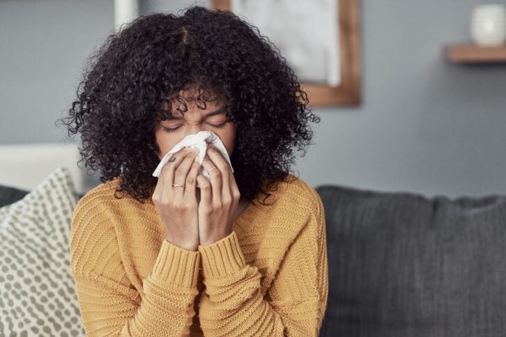 Midsection and head of young woman in the midst of blowing her nose with a tissue while sitting on a dark grey couch at home, with a blurred background of grey wall