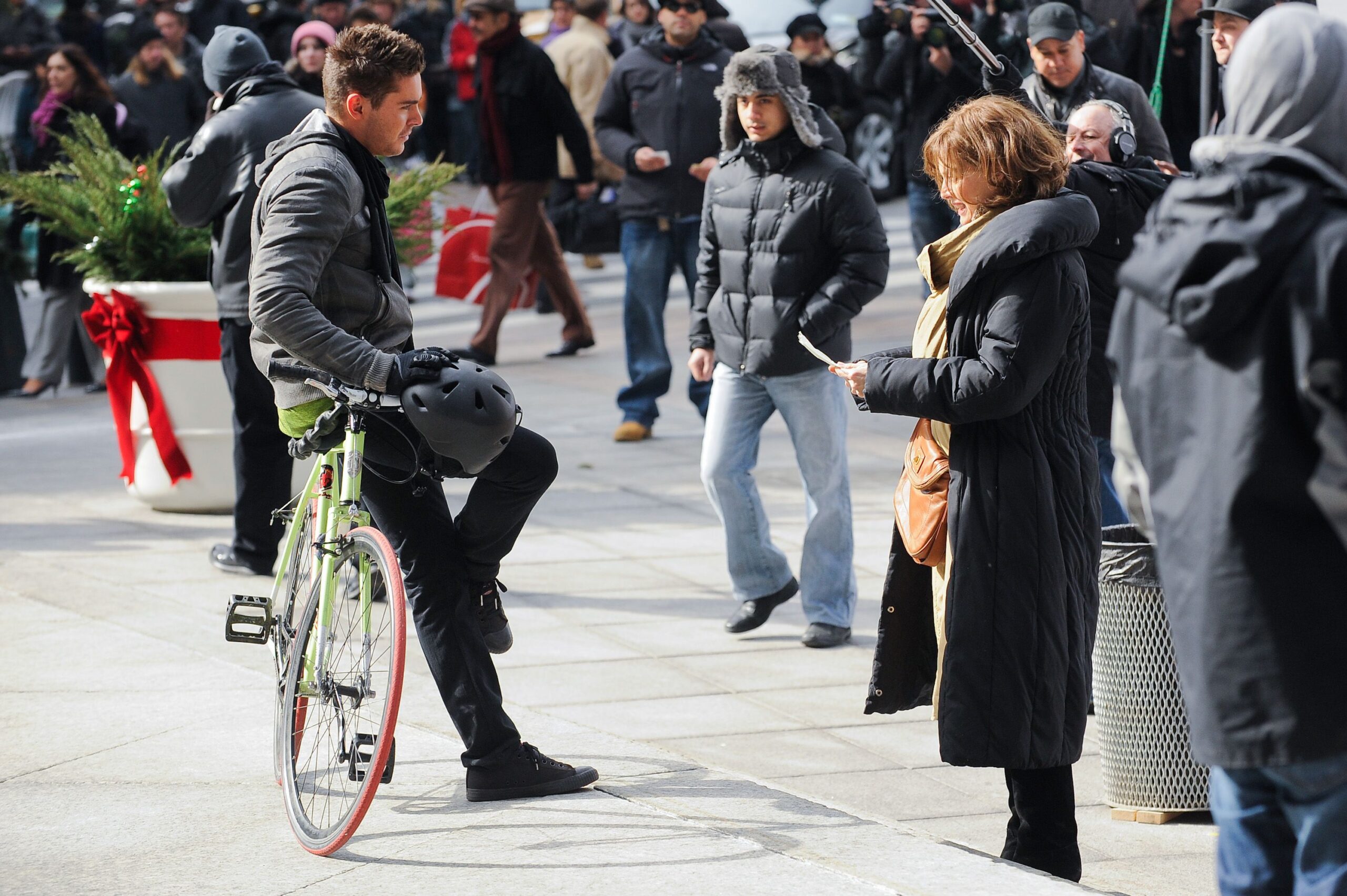 A man with a bicycle talks to a woman holding a piece of paper on a busy city sidewalk. People in winter clothing walk by, and holiday decorations are visible in the background.