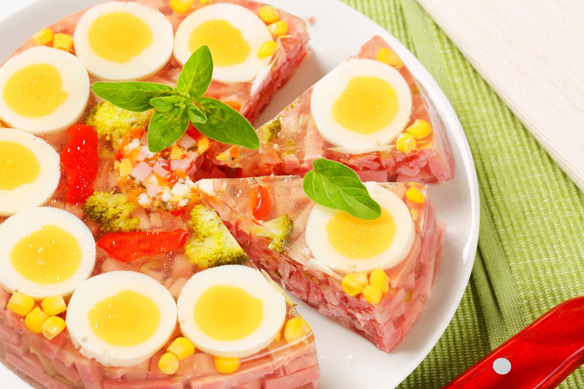 A colorful gelatin salad with slices of boiled egg, corn, broccoli, red pepper, and diced ham, garnished with fresh basil leaves, served on a white plate.