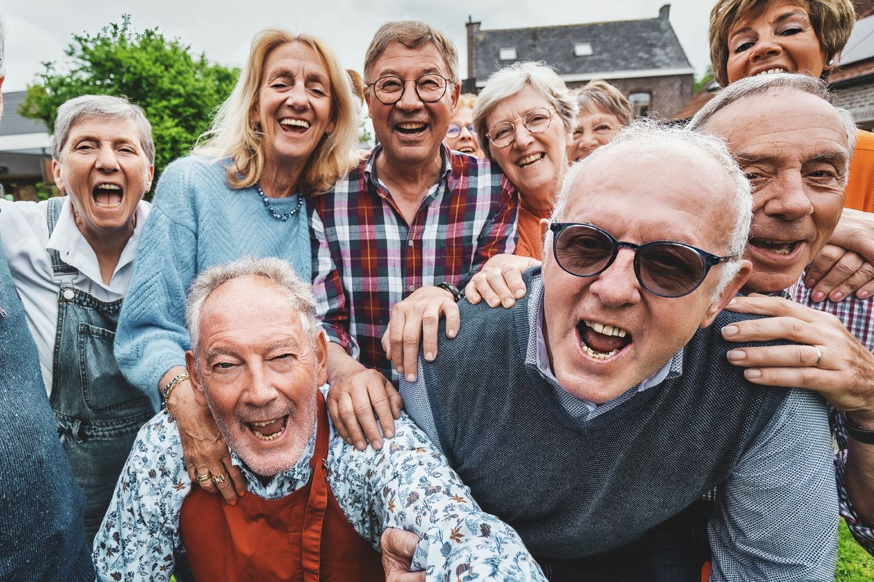 A group of happy older adults gather closely outdoors, smiling and laughing at the camera on a bright day, suggesting a joyful, social occasion. Some have their arms around each other. Trees and houses are in the background.