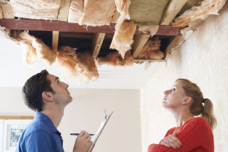 repairman with clipboard inspecting ceiling damage while concerned woman talks on phone
