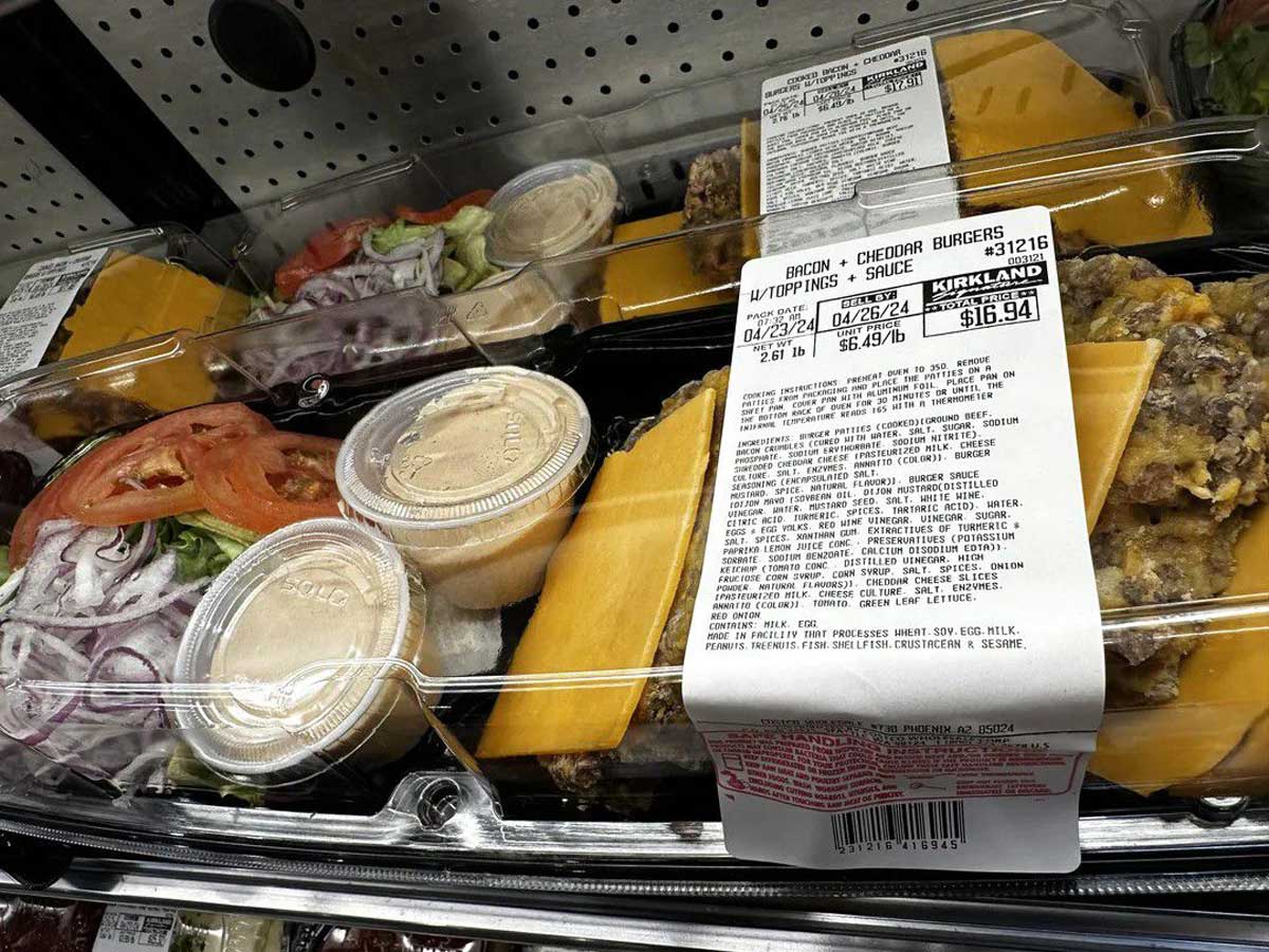 A plastic container of Kirkland bacon cheddar burgers with cheese slices, sliced onions, tomatoes, lettuce, and sauce cups—part of the Costco meal kits—displayed on a store shelf with a price label visible on top.