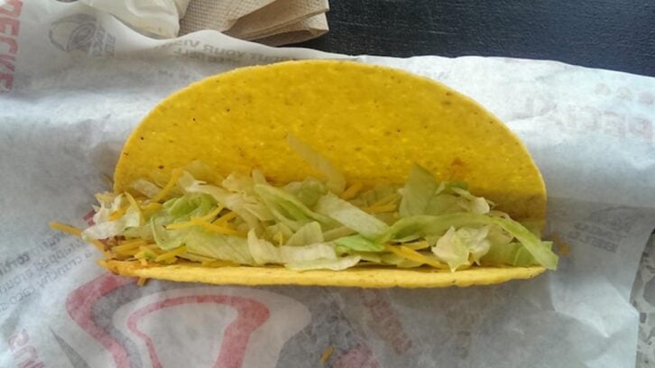 A hard shell taco filled with shredded lettuce and shredded cheese sits on a wrapper with some napkins in the background.