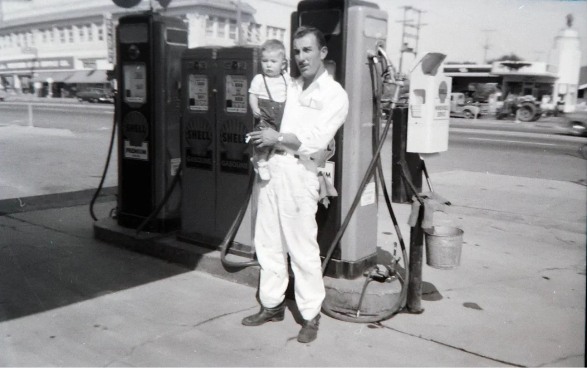 A man in a white jumpsuit holds a small child while standing next to vintage Shell gas pumps at an old-fashioned gas station on a sunny day.