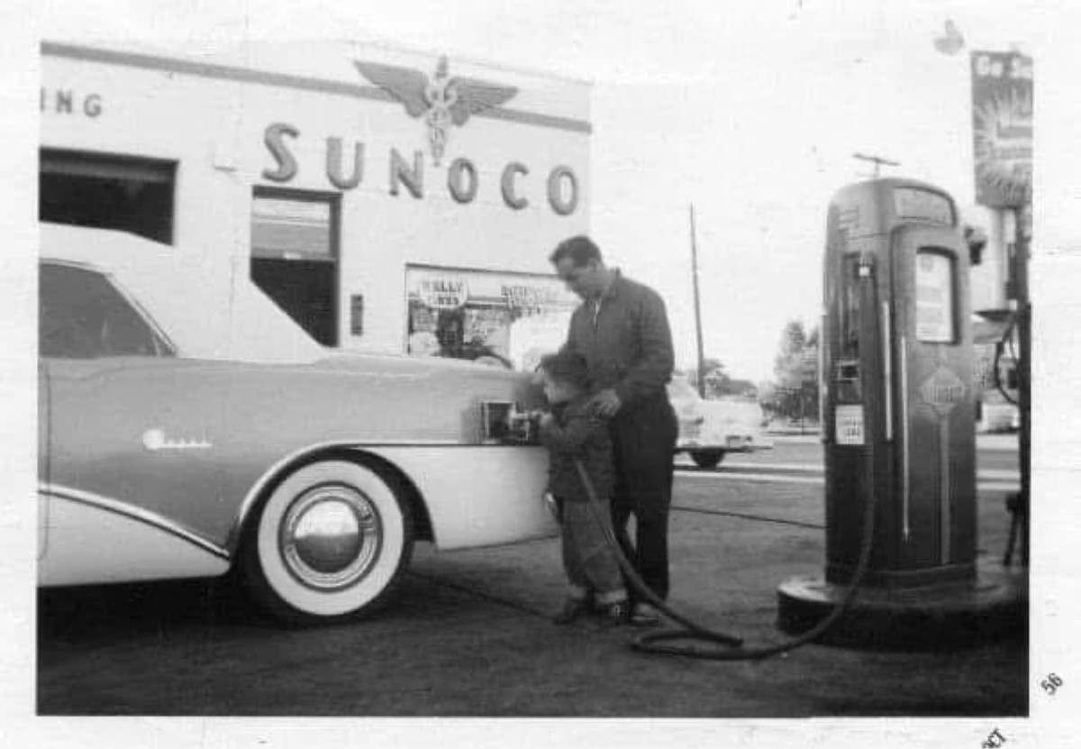 A man helps a young boy pump gas into a classic car at a Sunoco gas station. An old-fashioned gas pump stands in the foreground, and other vintage cars are visible in the background. The scene appears to be from the 1950s.