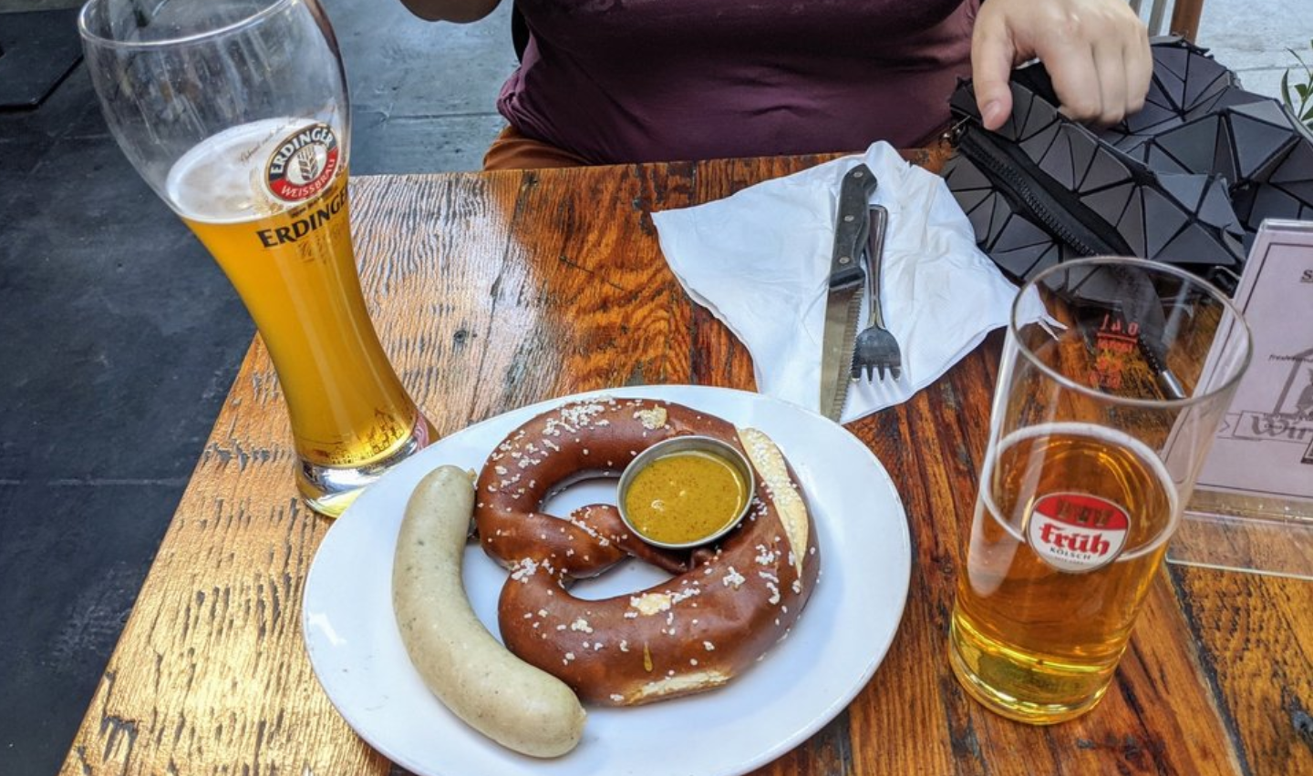 A wooden table with a white plate holding a large pretzel, a white sausage, and mustard. Next to it are two glasses of beer, and a person is seated in the background with a hand near a black purse.