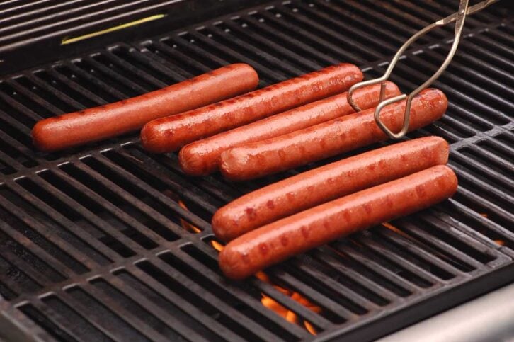 Six hot dogs are being grilled on a barbecue; five are lying on the grill, and one is being turned with metal tongs. There are visible flames and grill marks on the sausages.