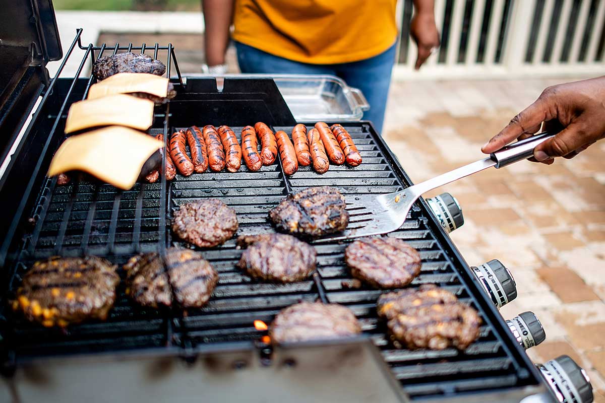 A person grilling hamburgers, sausages, and burger buns with cheese on an outdoor barbecue grill, with another person standing nearby in the background.