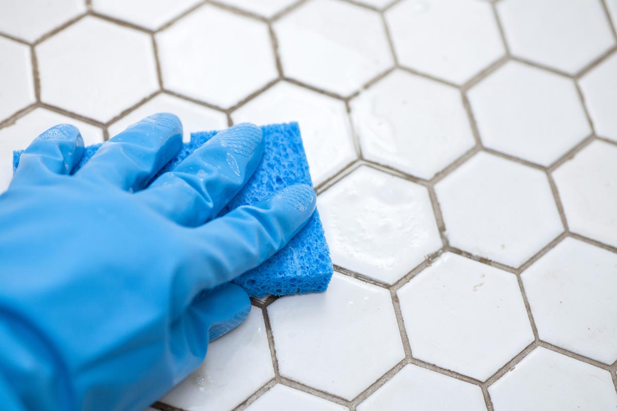 A hand wearing a blue glove is scrubbing white hexagonal tile with a blue sponge, cleaning the grout between the tiles.