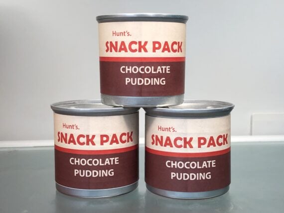 Three cans of Hunt’s Snack Pack chocolate pudding are stacked on a smooth surface, with one can on top of two others. The labels are beige and brown with red and white text.