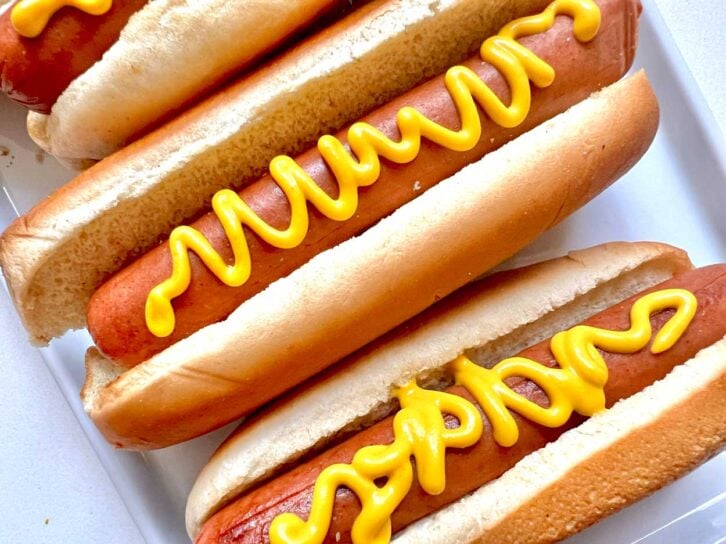 Three hot dogs in buns, each topped with a squiggly line of yellow mustard, are arranged on a white plate.