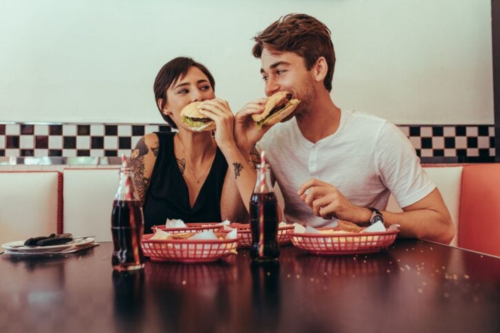 Romantic couple sitting at a diner and eating burgers interlocking their arms looking at each other. Couple at a restaurant eating burgers with soft drinks on the table.