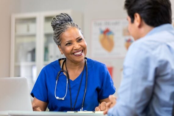 a cheerful senior female nurse sits at a table with an unrecognizable patient