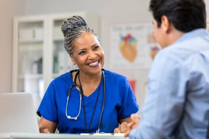 a cheerful senior female nurse sits at a table with an unrecognizable patient