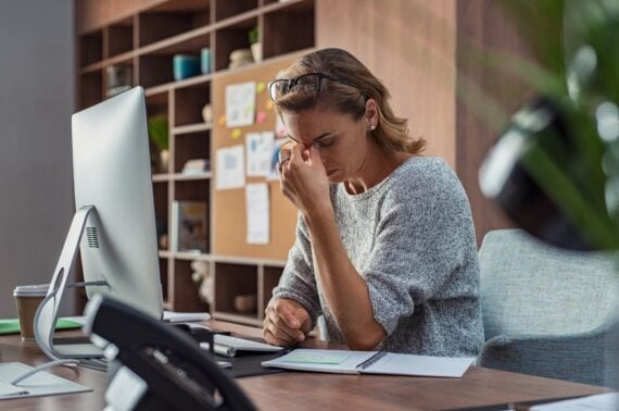 Exhausted businesswoman having a headache in modern office. Mature creative woman working at office desk with spectacles on head feeling tired. Stressed casual business woman feeling eye pain while overworking on desktop computer.