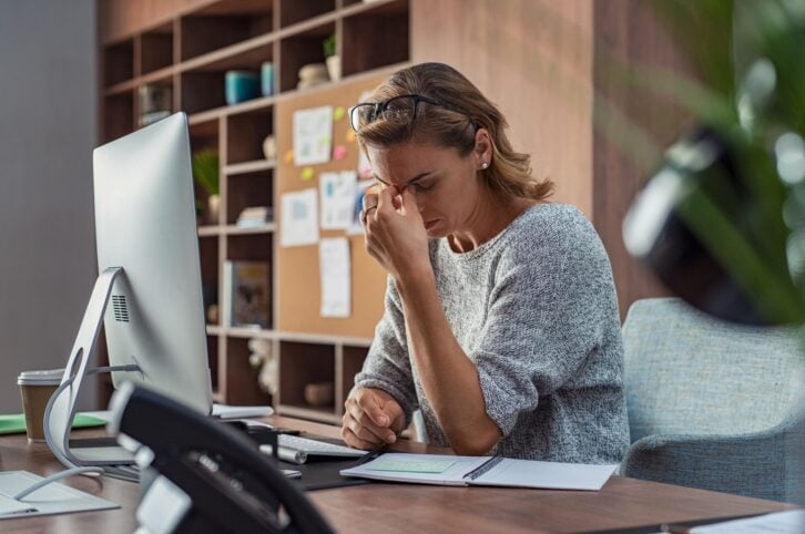 Exhausted businesswoman having a headache in modern office. Mature creative woman working at office desk with spectacles on head feeling tired. Stressed casual business woman feeling eye pain while overworking on desktop computer.