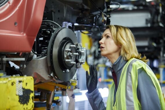 Confident mature female maintenance engineer examining car at factory. Blond manager is checking vehicle at automobile industry. She is wearing reflective clothing.
