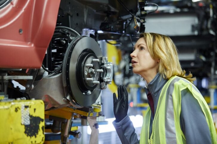 Confident mature female maintenance engineer examining car at factory. Blond manager is checking vehicle at automobile industry. She is wearing reflective clothing.