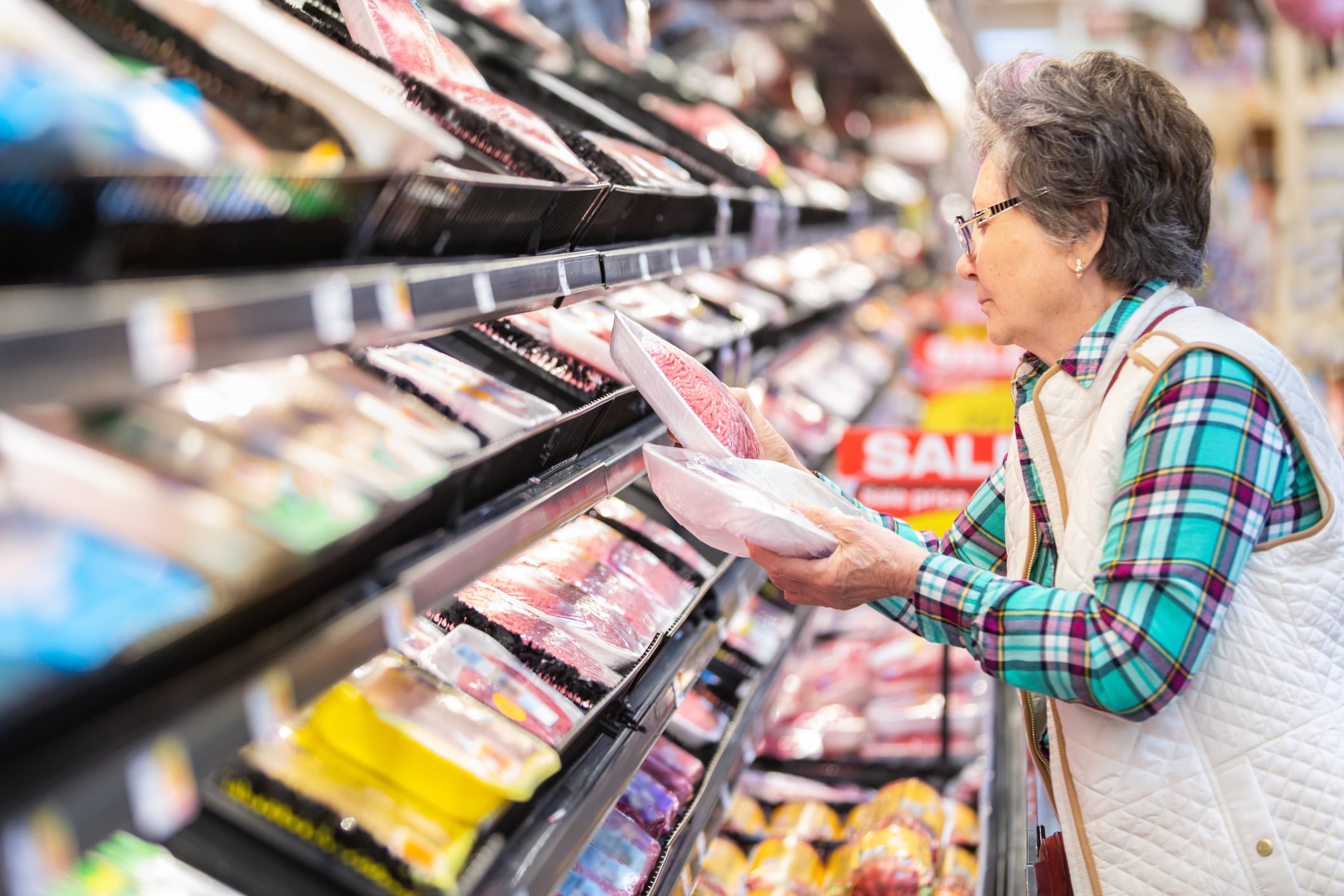 An older woman wearing glasses and a plaid shirt examines packaged meat in a grocery store aisle, surrounded by various meat products, with a "SALE" sign visible in the background.
