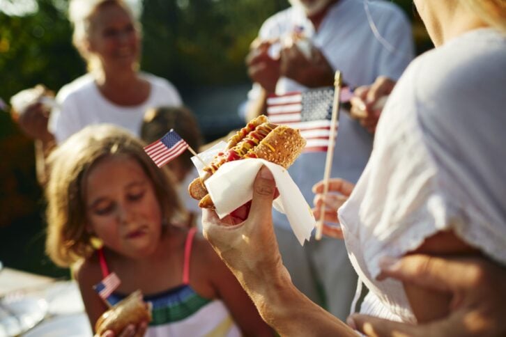 Multi-generation family on picnic in back yard, eating hot dog and celebrating 4th of July - Independence Day. Focus on hot dog with USA flag.