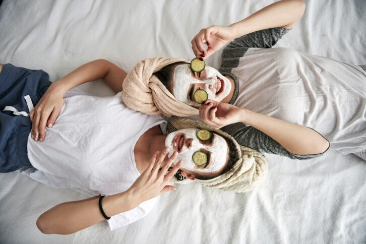 two people with face masks and cucumber slices on their eyes laying on bed