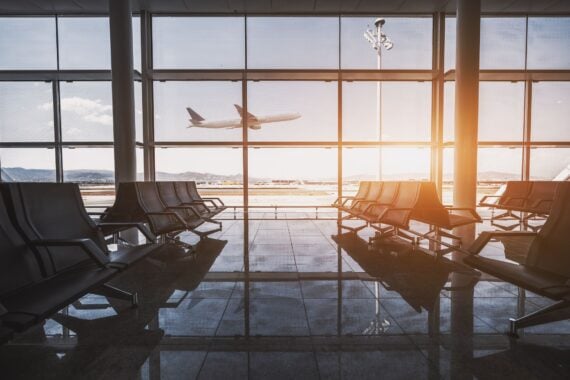 Wide-angle view of a modern aircraft gaining the altitude outside the glass window facade of a contemporary waiting hall with multiple rows of seats and reflections indoors of an airport terminal El Prat in Barcelona