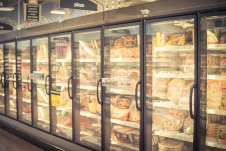 Blurred variety pack of processed pizza at retail store in America. Frozen food section in huge glass door aisle. Freezer full assortment of frozen pizza in local supermarket, defocused background