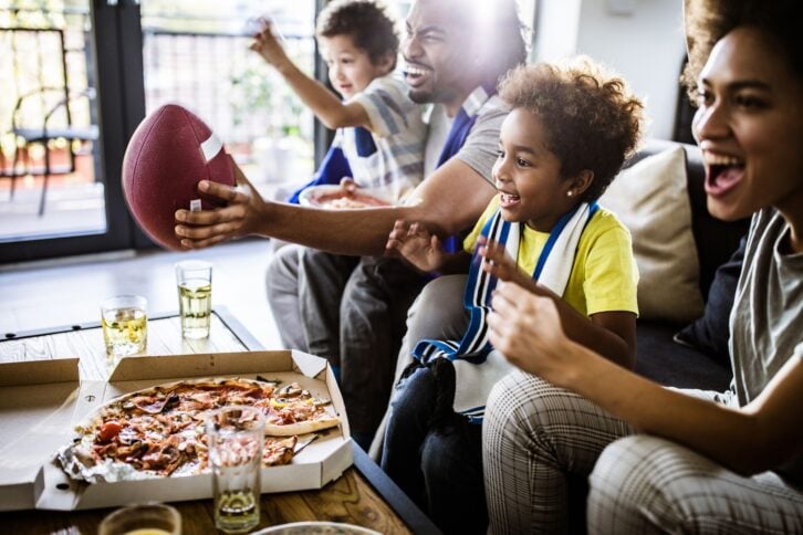 Young joyful family eating pizza having fun while cheering for their favorite American football team at home