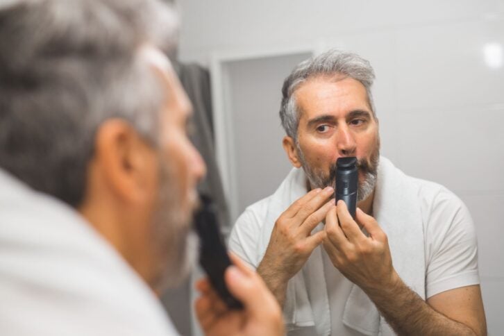 middle aged man trimming his beard in bathroom with electric shaver
