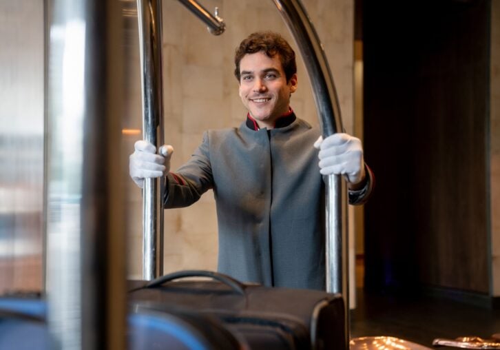Young bellboy at a hotel moving luggage cart to a guest room very cheerfully and smiling