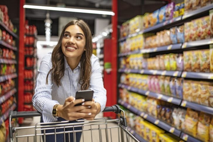 Smiling woman at supermarket. Happy woman at supermarket. Beautiful young woman shopping in a grocery store/supermarket. Shopping lists in app format
