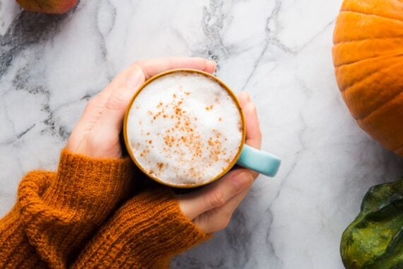 Autumn moody background with mug of latte coffee and pumpkins on marble table. Flat lay in fall colors. Female hands in cozy sweater