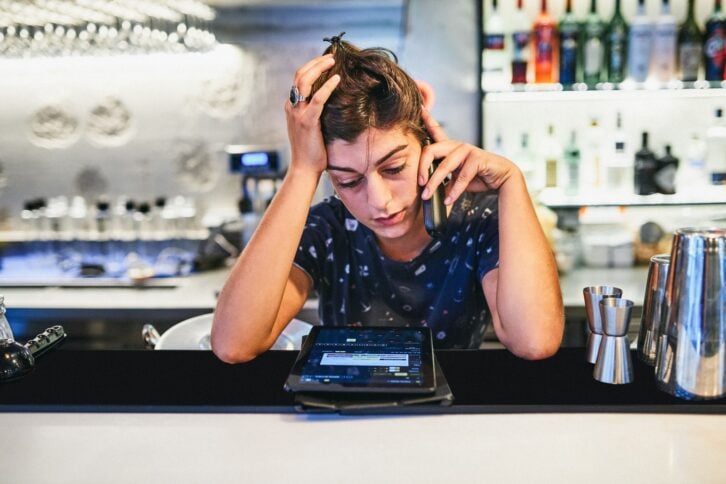 Shot of a young woman looking stressed while using a phone behind the counter of a bar