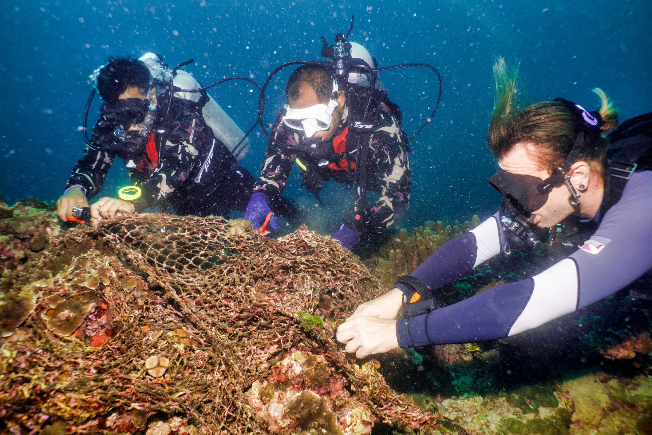 three scuba divers are cutting discarded fishing net from an underwater coral reef in thailand