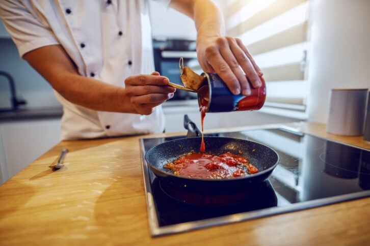 Close up of creative experienced caucasian chef in uniform adding peeled tomato. Domestic kitchen interior.