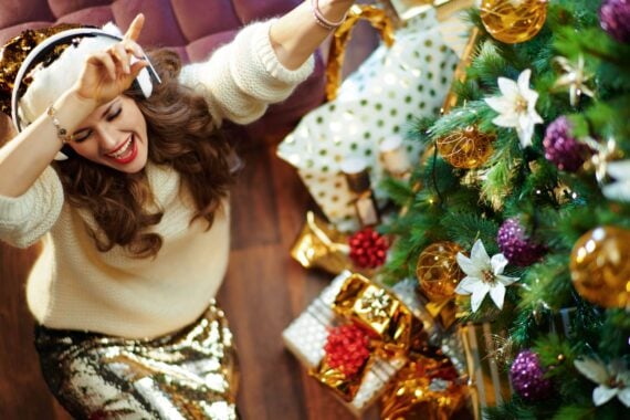Upper view of happy trendy woman with long brunette hair in gold sequin skirt and white sweater listening to the music with headphones and dancing under decorated Christmas tree near present boxes.