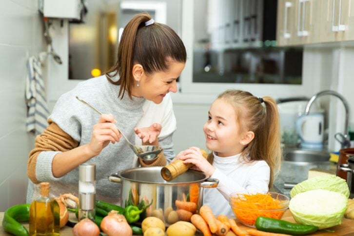 Positive mother and little daughter tasting soup together at home