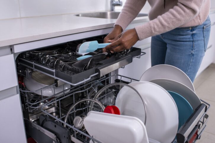 Close up image of a woman's hands holding a spatula, taking out clean cutlery from the dishwasher rack.