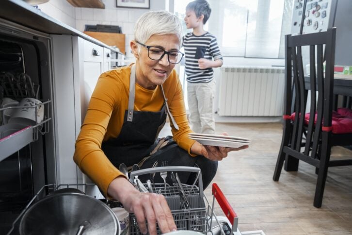 Grandma with boy washing dishes together in kitchen , taking out dishes from dishwashing machine