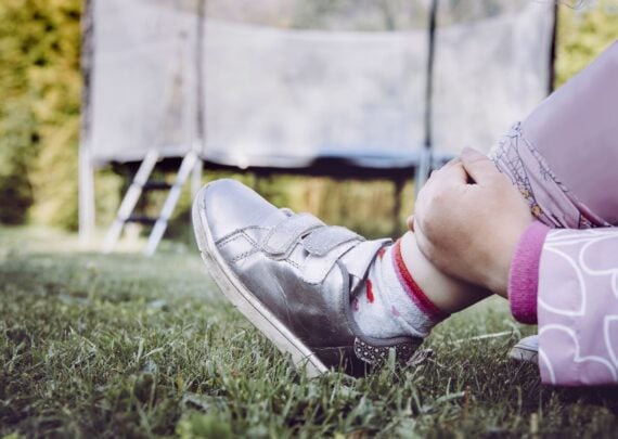 Trampoline jumping accident concept. Child holding broken sprained ankle with hands outdoors by the home garden trampoline.