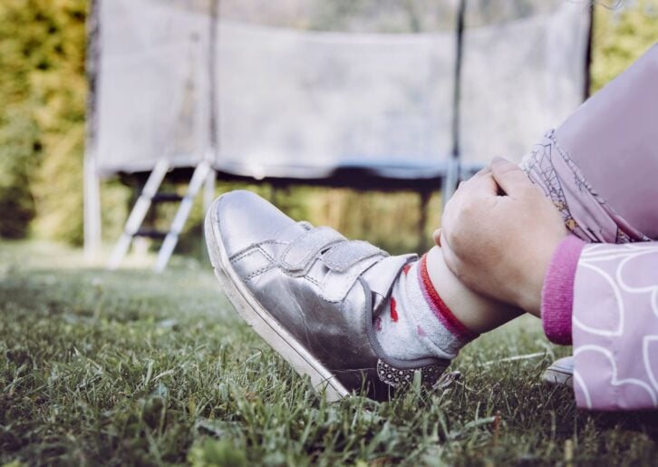 Trampoline jumping accident concept. Child holding broken sprained ankle with hands outdoors by the home garden trampoline.