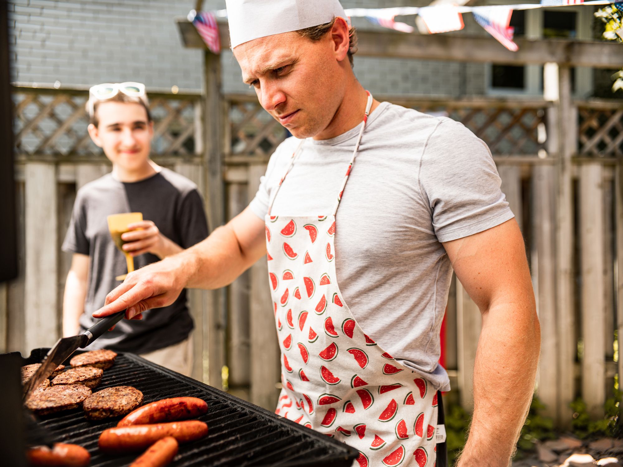 Young Caucasian man in company of boy teenager setting up a BBQ grill for outdoor 4th July backyard celebration.