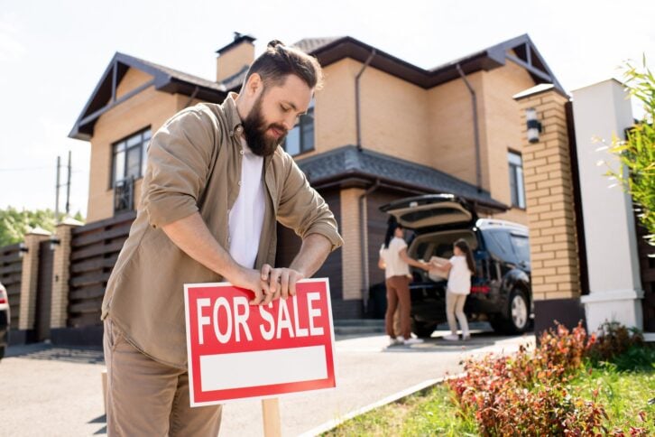 Young bearded father placing For sale sign into ground near house while putting house up for sale