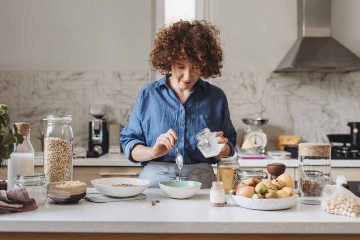 Homemade baked granola: woman adding ingredients into a bowl of rolled oats in the kitchen.