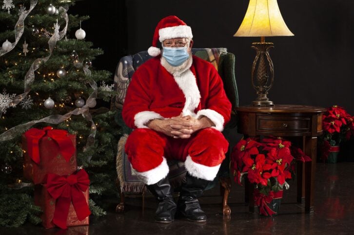 horizontal photograph of Santa Claus Sitting in a Chair Wearing a Surgical Mask and Looking Towards the Camera