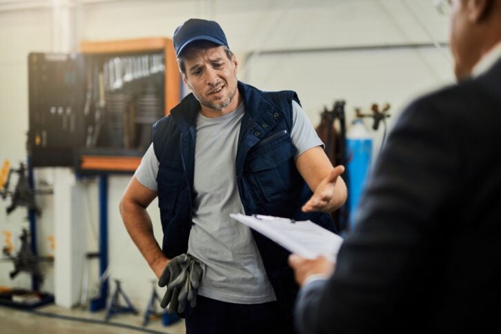 Auto repairman feeling uncertain while discussing about reports with his manager in a workshop.