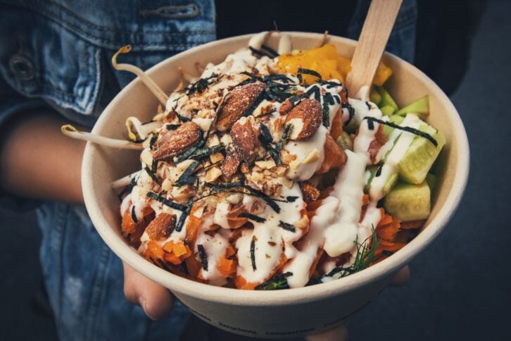 Disposable cardboard bowl with fresh vegetarian salad in hand of a young woman on the street. Every day more and more restaurants offer vegetarian salads as a healthy take out food option.