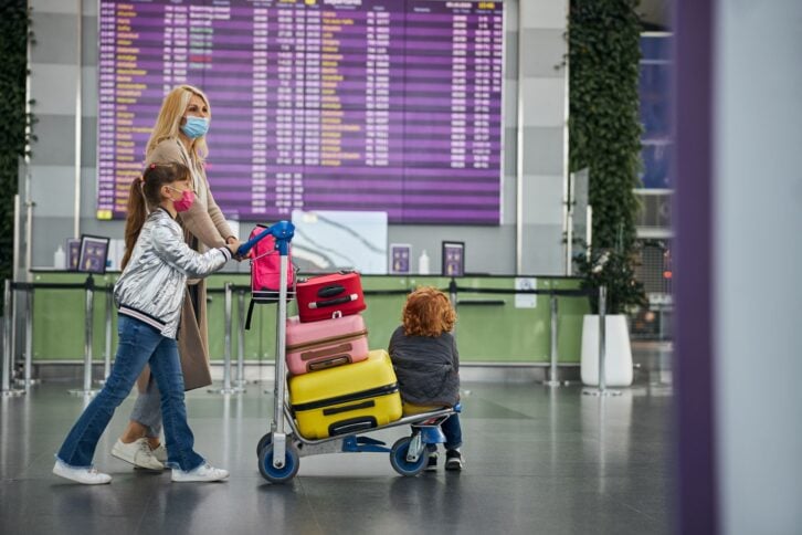 Girl and her mother wearing surgical masks rolling a trolley with a baggage and a boy past an information board