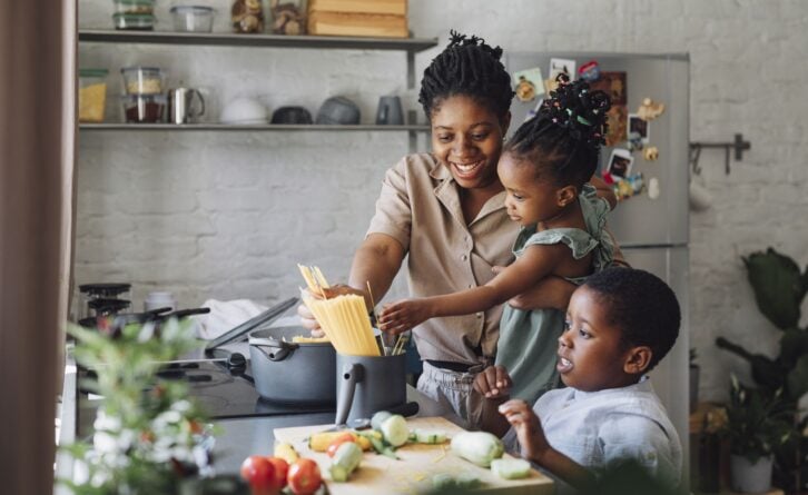 Happy African American mother, daughter and son preparing spaghetti and vegetables for lunch over a kitchen sink.