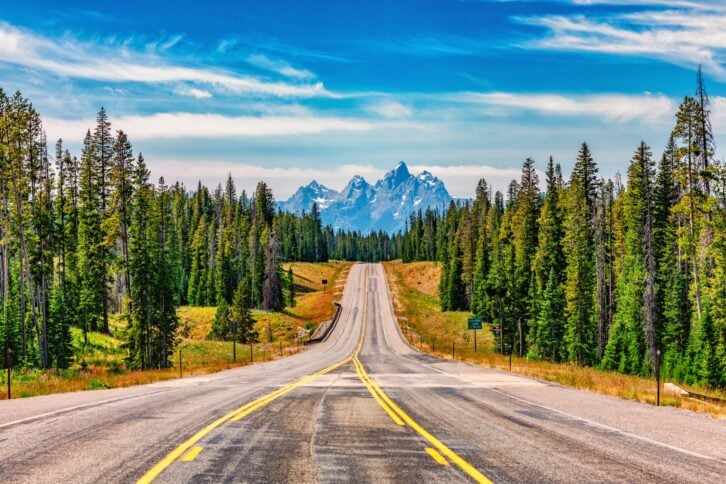 A mountain highway through the forest toward Grand Teton National Park, Wyoming with the peaks of the Tetons in the background.